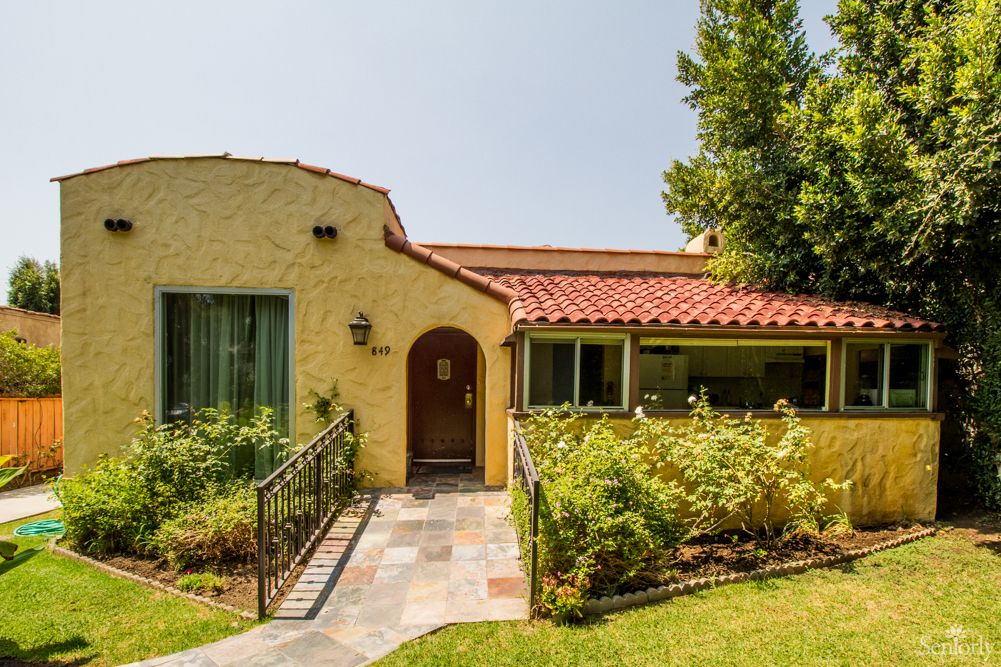 A charming yellow house with a red-tiled roof, surrounded by a lush garden and a neatly paved pathway.