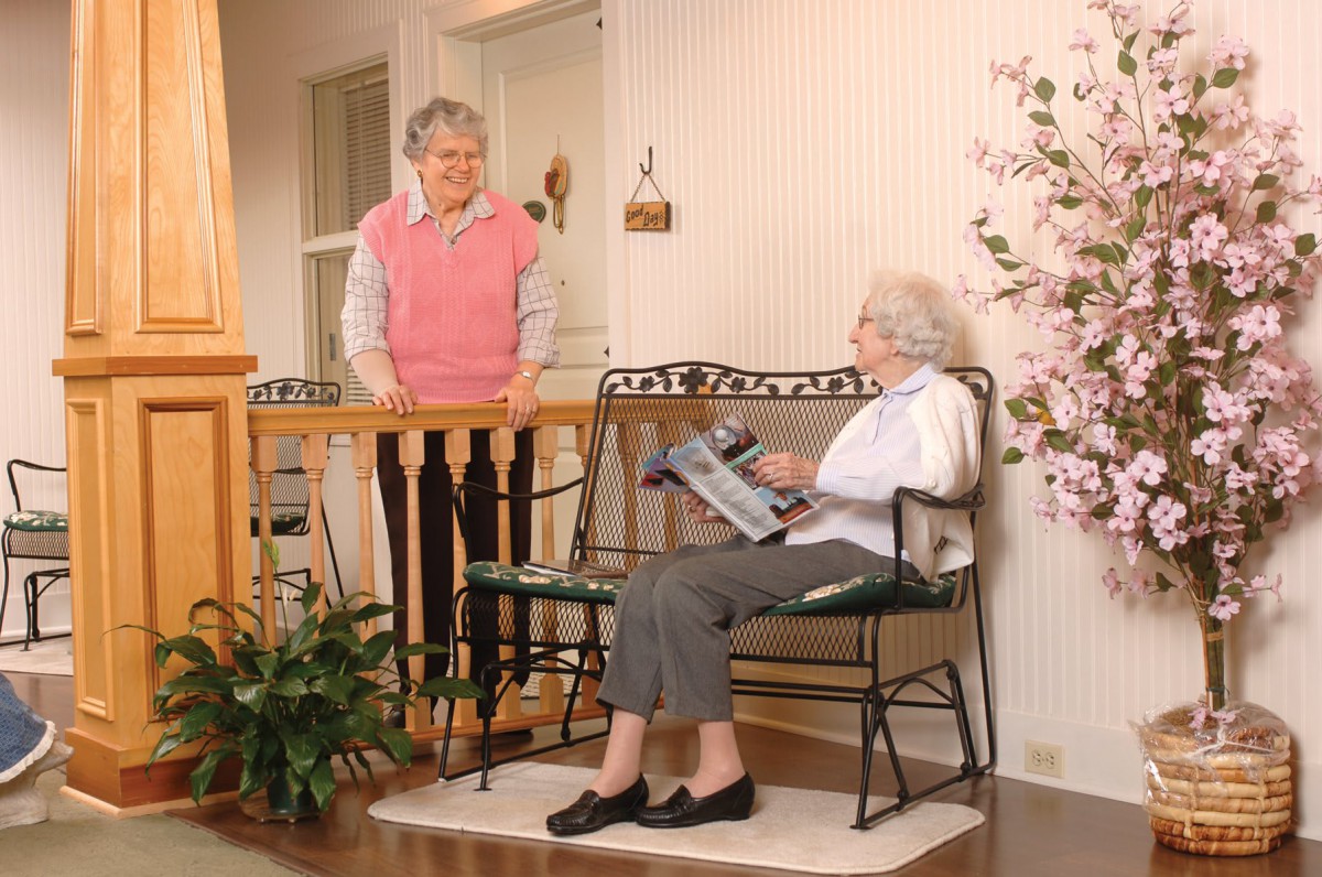 Two elderly women conversing in a cozy home setting, with one sitting and reading a magazine.