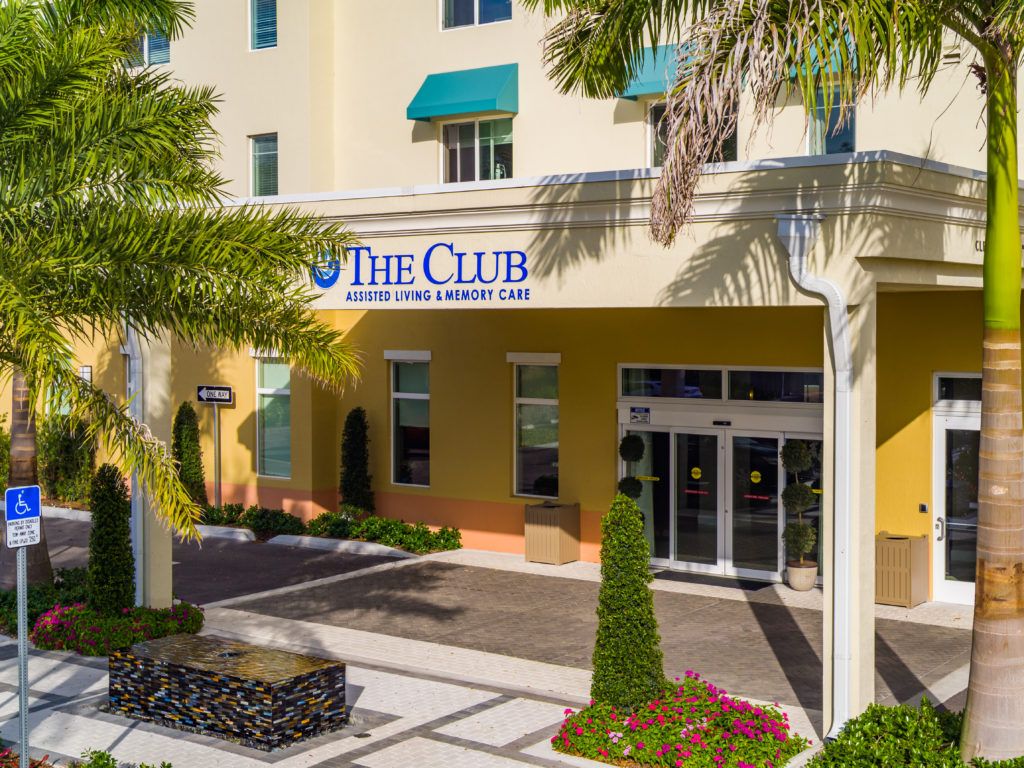 Entrance to a building with 'The Club' sign and surrounding palm trees and landscaping.