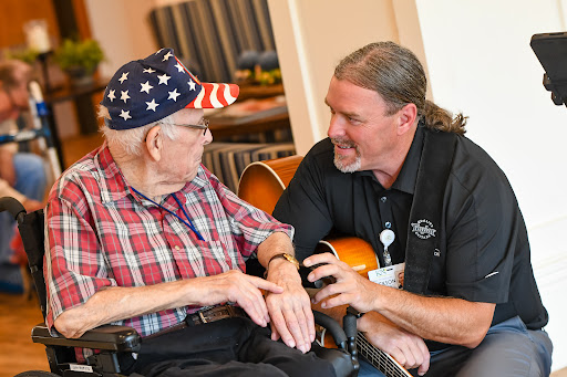 An elderly man wearing a patriotic cap shares a smile with a caregiver while enjoying a moment of music together.