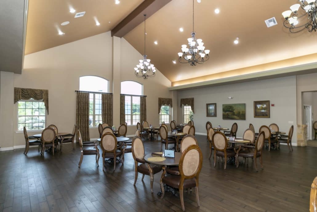 A spacious dining area with elegant tables and chairs, illuminated by chandeliers and natural light from large windows.
