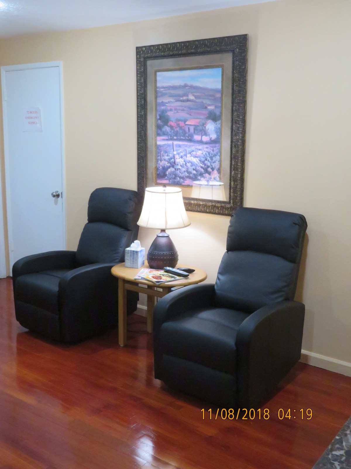 A cozy waiting area with two black recliner chairs, a wooden side table with a lamp, and a painting on the wall.