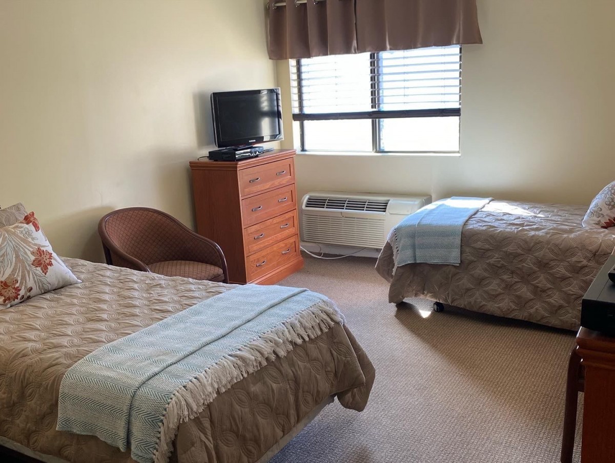 A hotel room featuring two twin beds, a television on a dresser, and a window with an air conditioner.