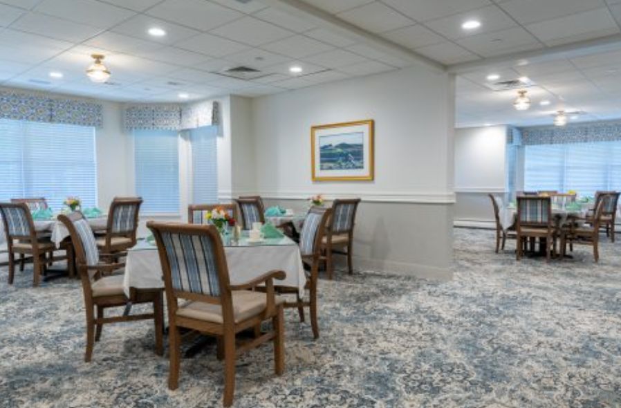A well-lit dining room with neatly arranged tables and chairs, featuring a patterned carpet and a painting on the wall.