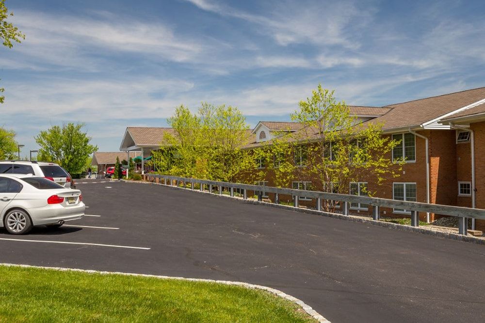 A spacious parking lot in front of a large building surrounded by trees under a blue sky with clouds.