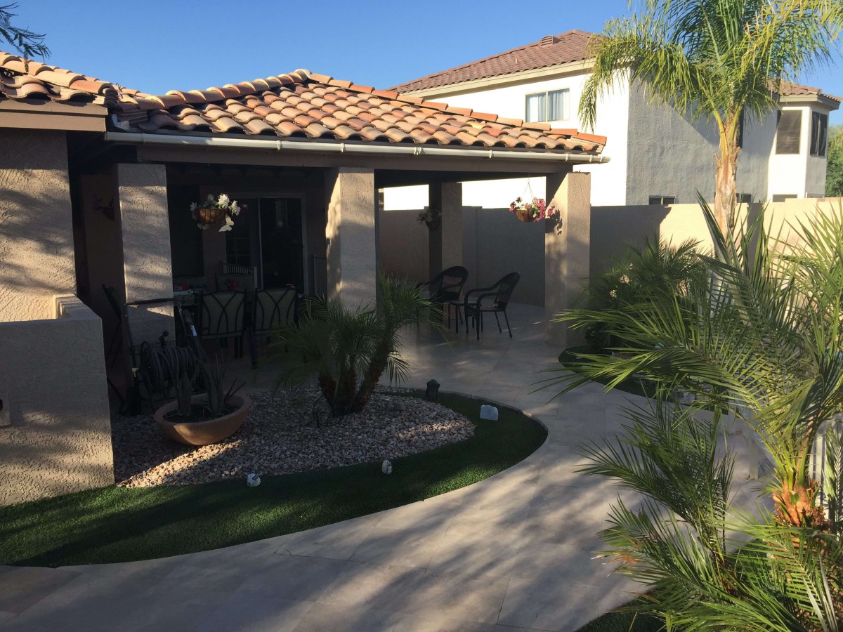 A cozy outdoor patio area with a terracotta tiled roof, surrounded by landscaping and palm trees.