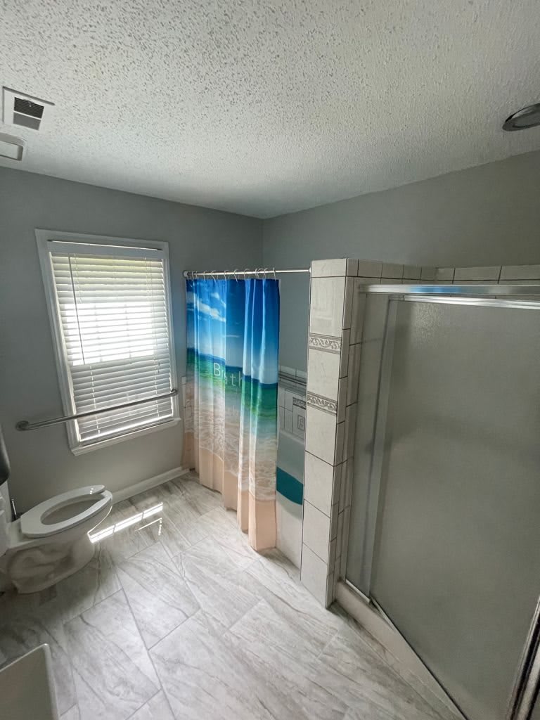 A bright and modern bathroom featuring a shower with a colorful curtain, a toilet, and a window with blinds.