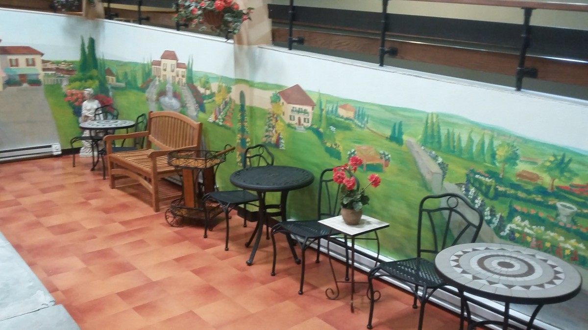 A cozy outdoor seating area featuring a mural of a countryside landscape, complete with tables, chairs, and decorative plants.