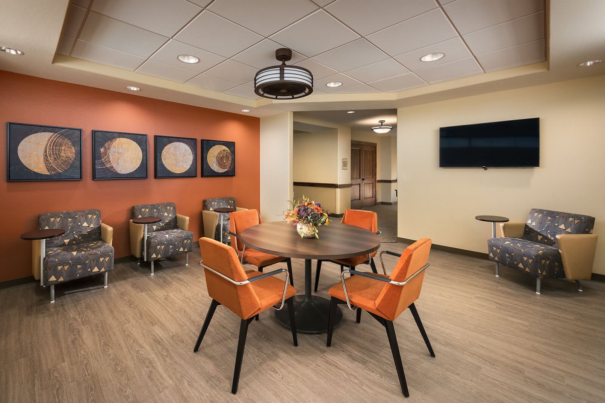 A modern lobby with comfortable seating, featuring a round table surrounded by orange chairs, wall art, and warm lighting.