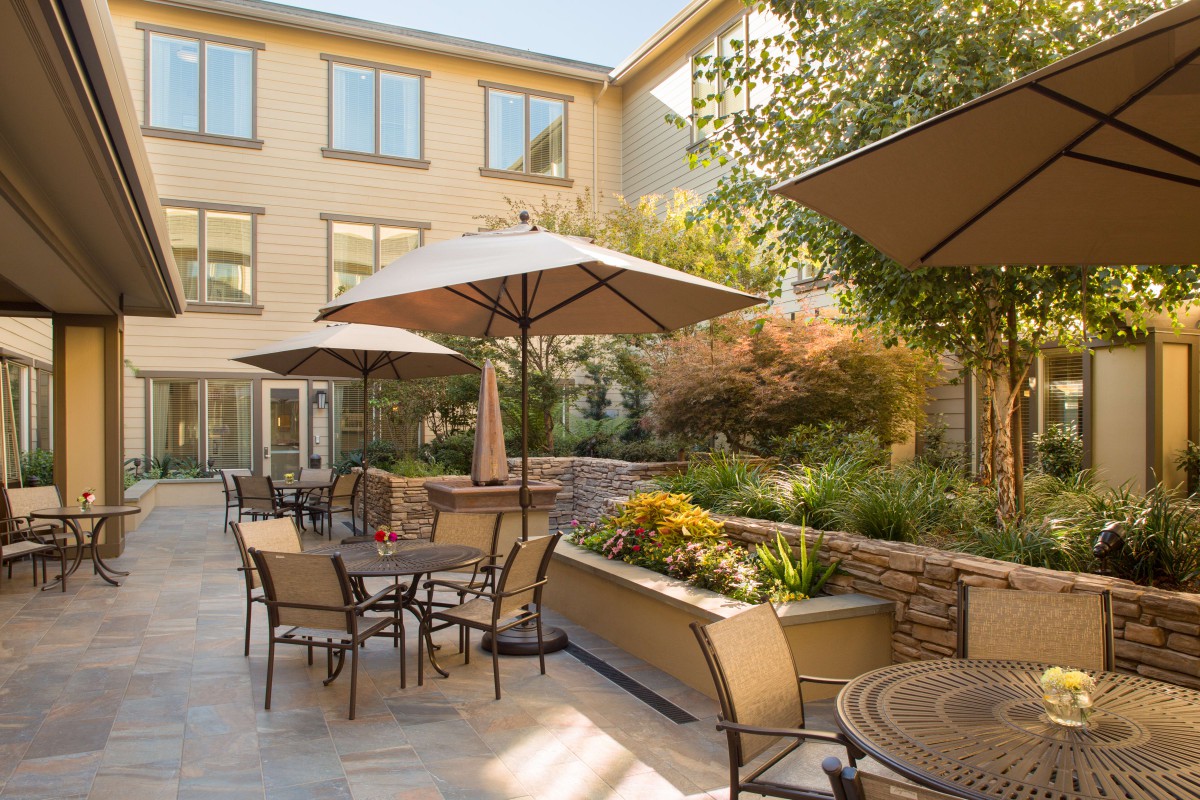 A sunny outdoor patio with tables, chairs, and umbrellas, surrounded by garden landscaping.
