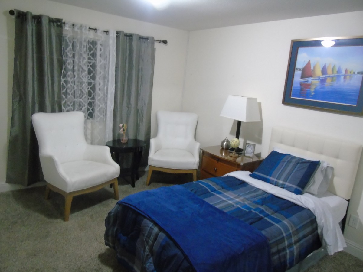 A cozy bedroom featuring a single bed with blue plaid bedding, two white armchairs, and a bedside lamp on a nightstand.