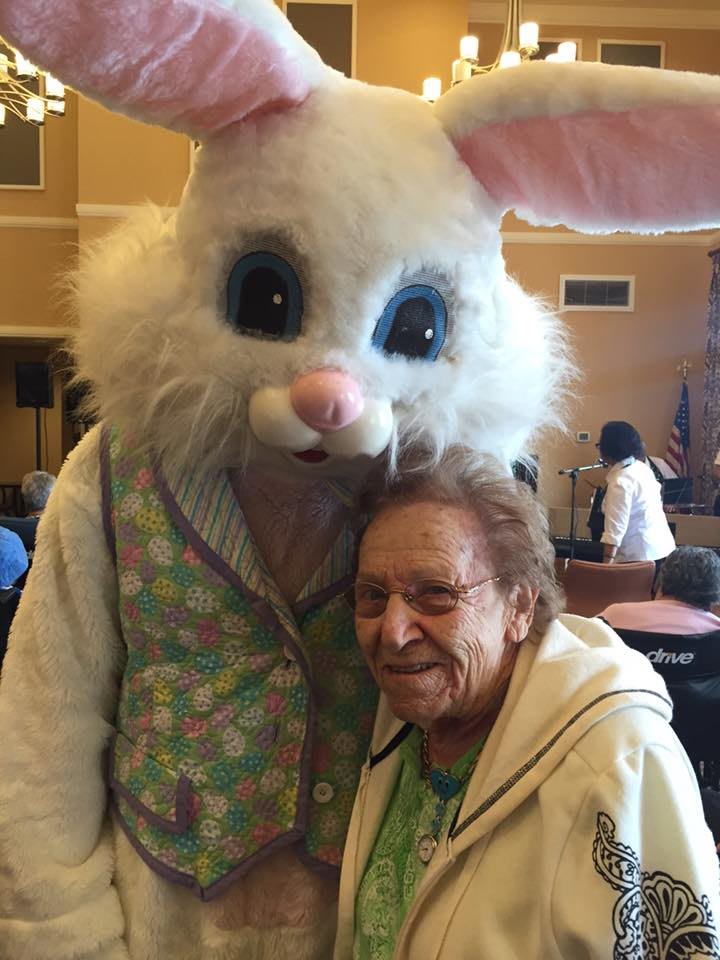 An elderly woman poses with someone dressed in a bunny costume, likely celebrating Easter indoors.