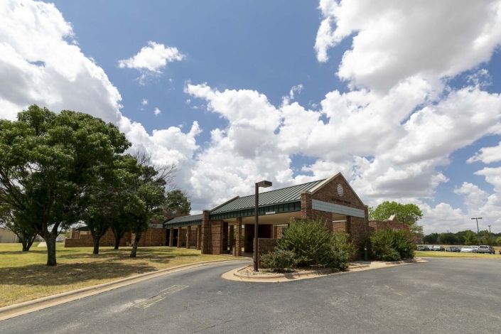 A brick building with a green roof under a partly cloudy sky, surrounded by trees and an asphalt driveway.