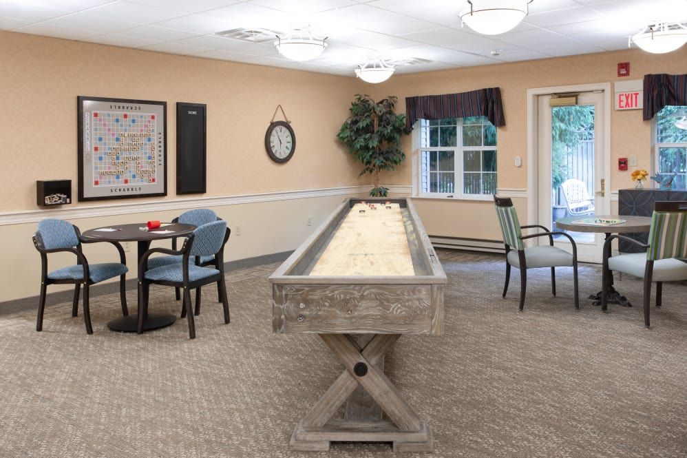 A cozy game room featuring a shuffleboard table, chairs, and a small round table set against carpeted flooring.