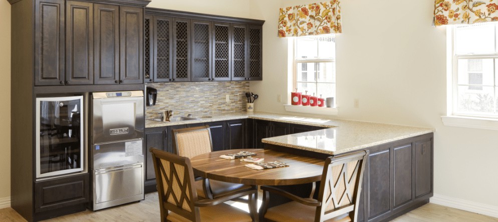 A cozy kitchen with a round wooden dining table, dark cabinets, and a small window with floral curtains.