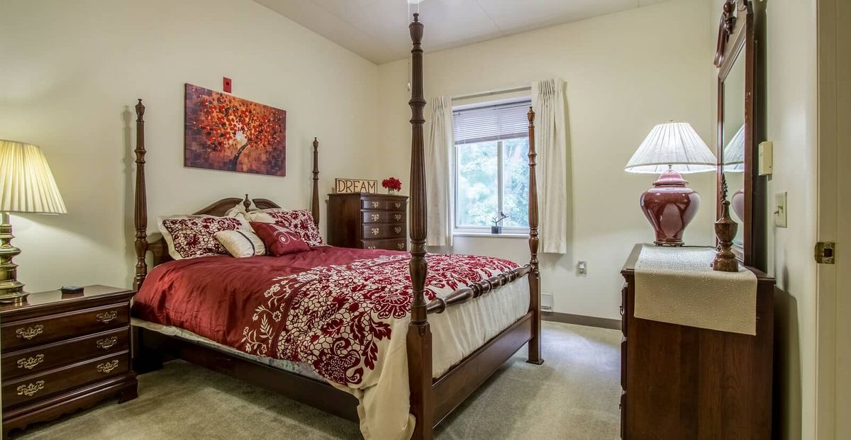 A cozy bedroom with a four-poster bed featuring red bedding, a window with curtains, and classic wooden furniture including a dresser and lamp.