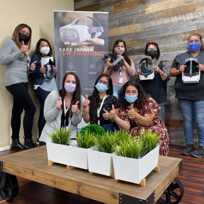 A group of people wearing masks and holding VR headsets pose for a photo indoors, with potted plants on a table.