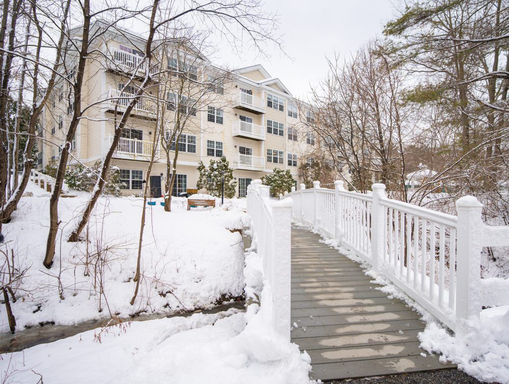 A snowy pathway leading to a multi-story apartment building surrounded by trees.