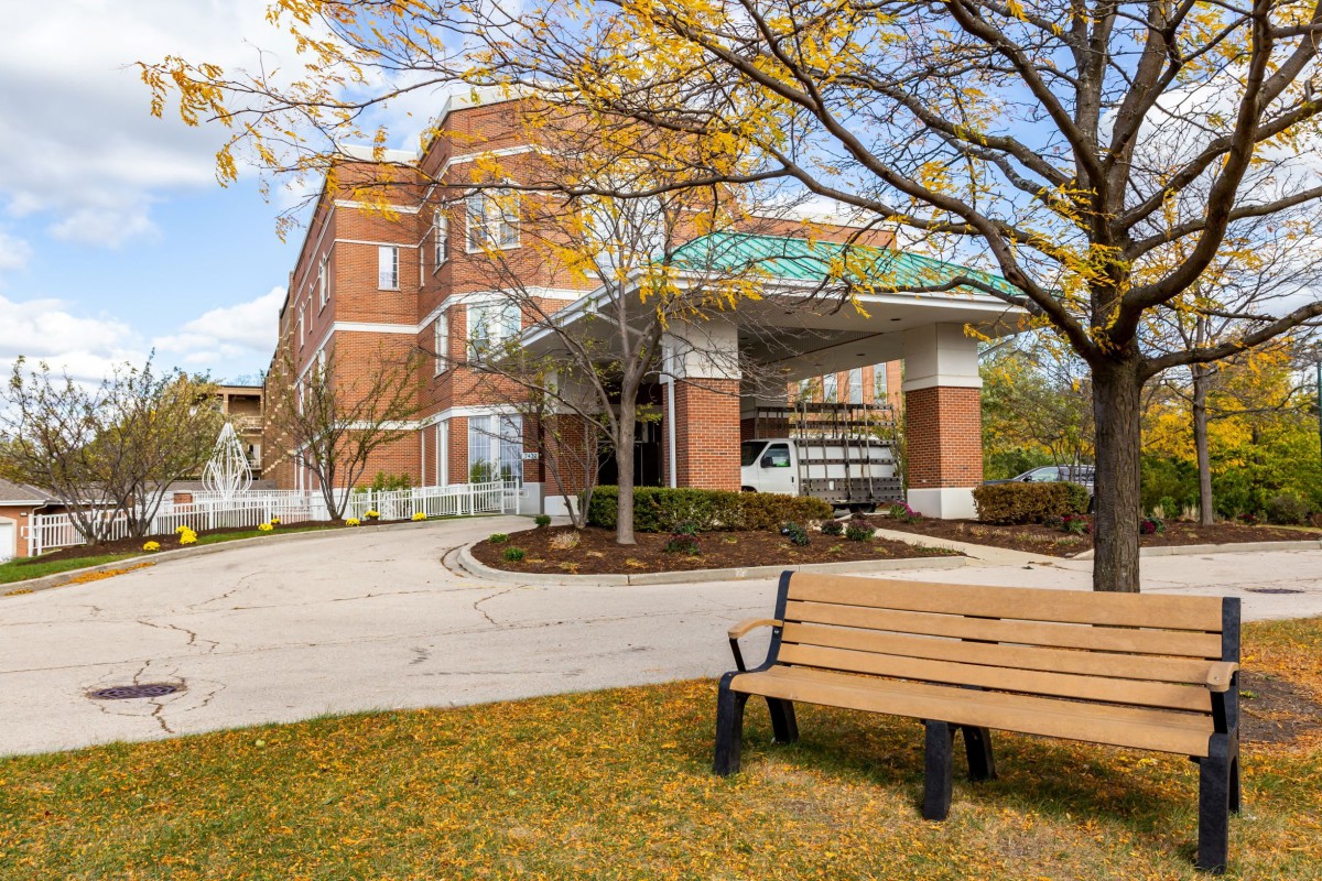 A brick hospital building with an entrance canopy, surrounded by autumn trees and a bench in the foreground.