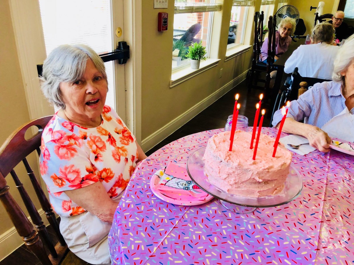 An elderly woman sitting at a table with a pink frosted cake and lit candles, celebrating a birthday.