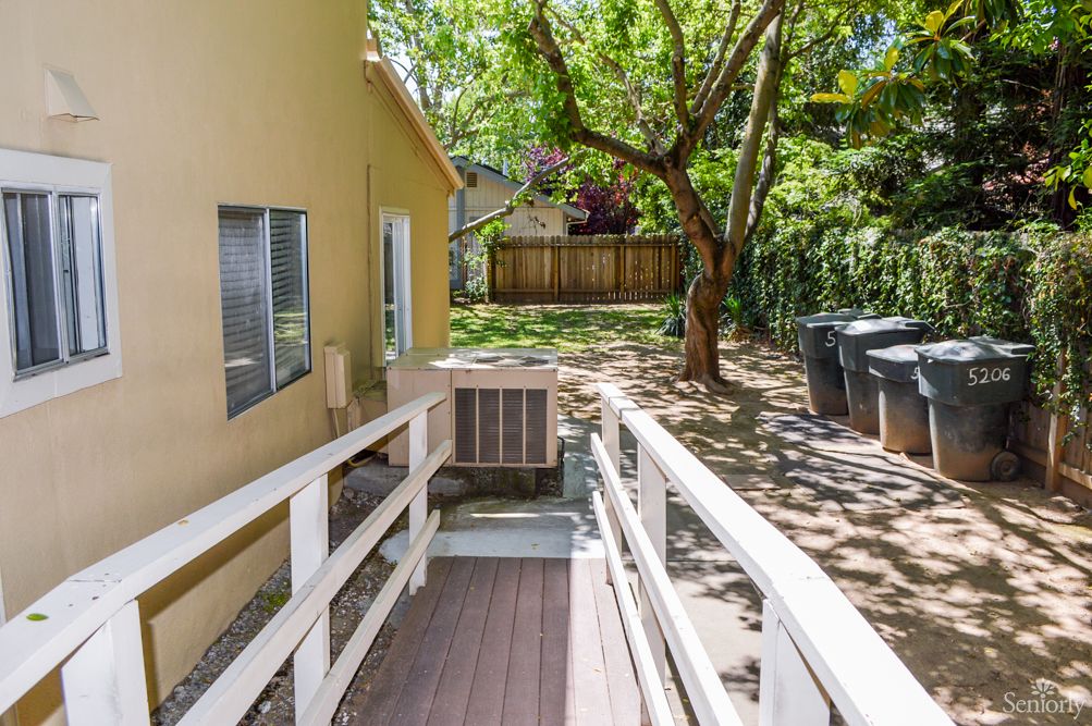A sunny backyard featuring a wooden ramp leading to a house, with a large tree providing shade.