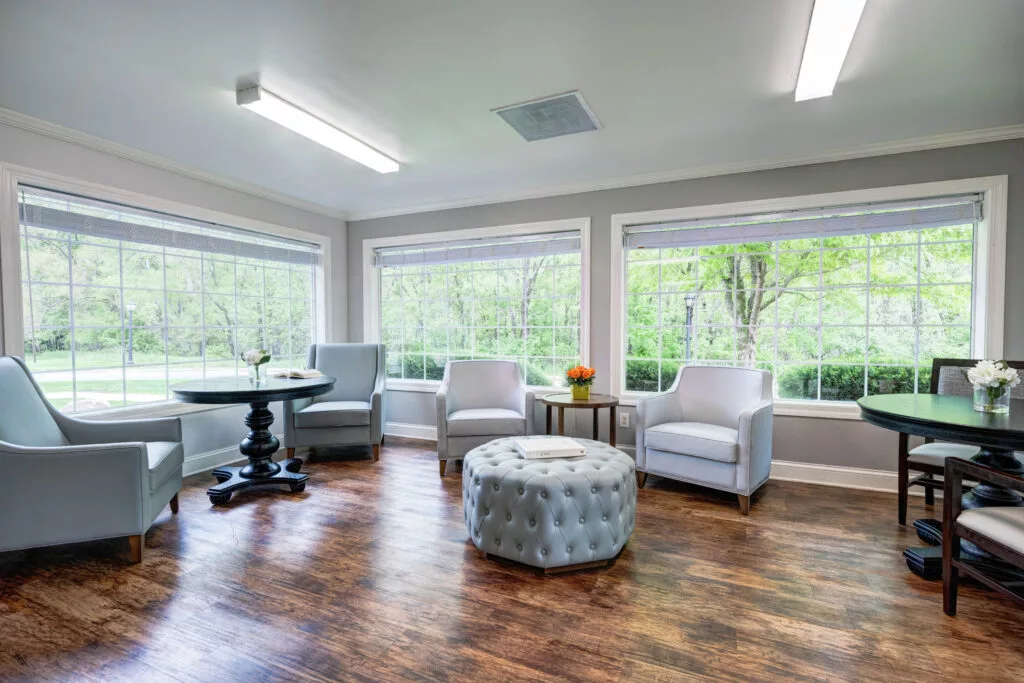 A cozy living room with ample natural light, featuring comfortable seating and a round ottoman on a hardwood floor.