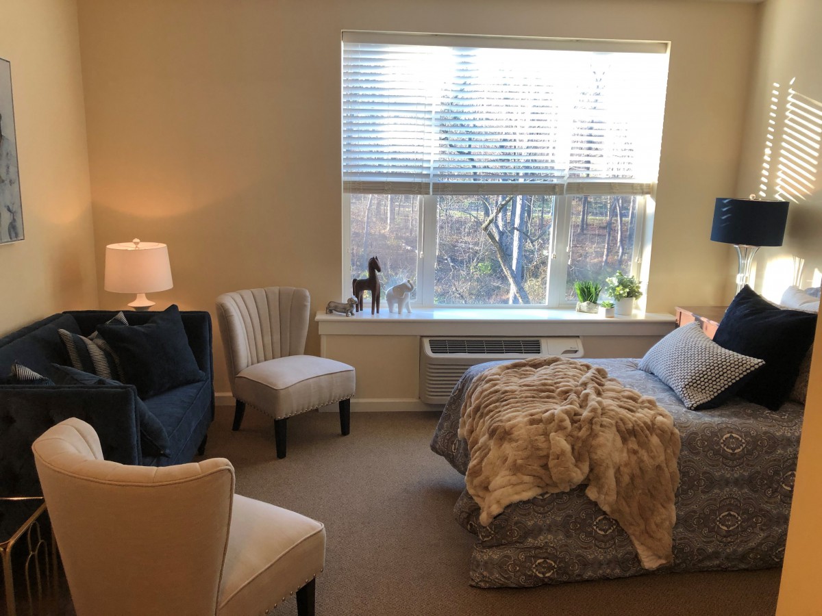 A cozy bedroom interior with a large window, bed, seating area, and soft lighting.