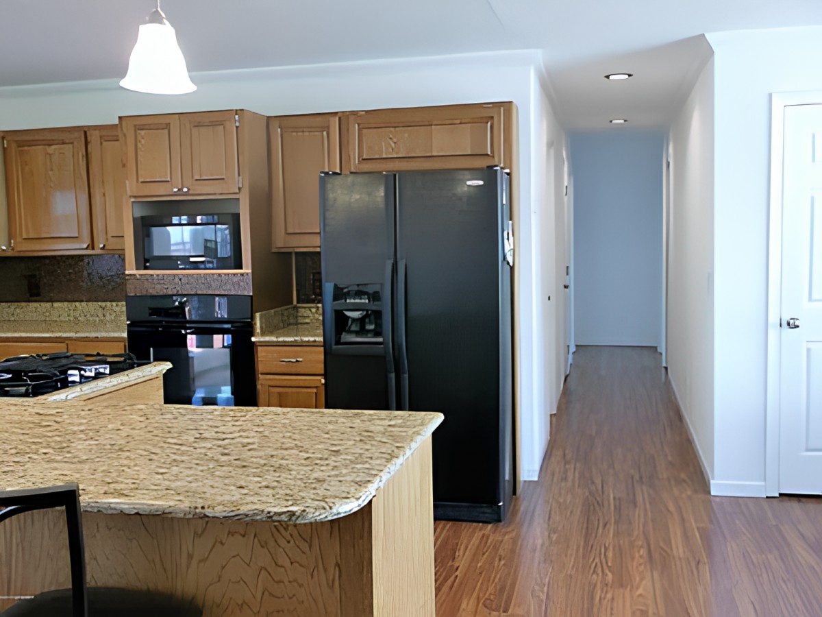 A modern kitchen with granite countertops, wood cabinets, and stainless steel appliances leading to a hallway.