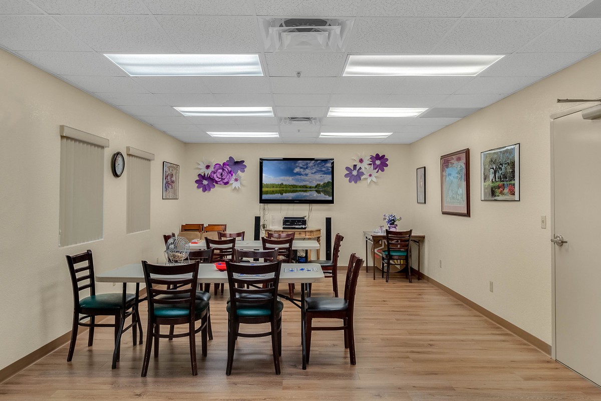 A well-lit communal room with tables, chairs, and a television mounted on the wall, decorated with artwork and purple floral designs.