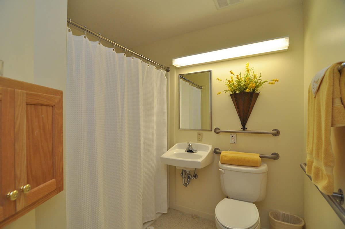 A neatly organized bathroom with a sink, toilet, and shower area. A mirror is mounted above the sink with decorative flowers and a towel placed on the toilet.