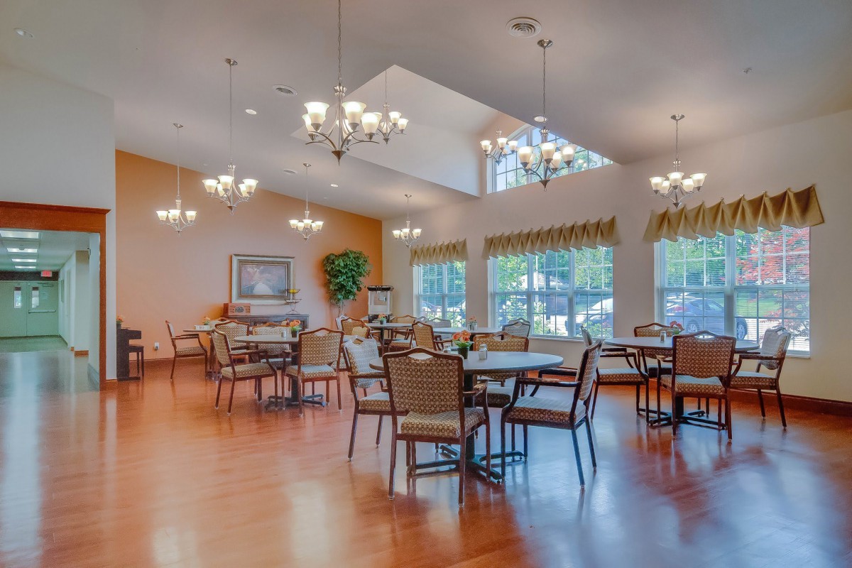 A bright and spacious dining room with multiple tables and chairs, featuring large windows and elegant chandeliers.