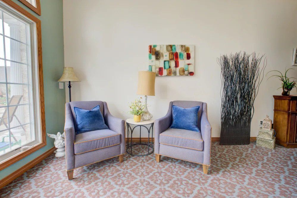 A cozy living room setup featuring two armchairs with blue throw pillows, a small round table with a lamp, artwork on the wall, and decorative plants.