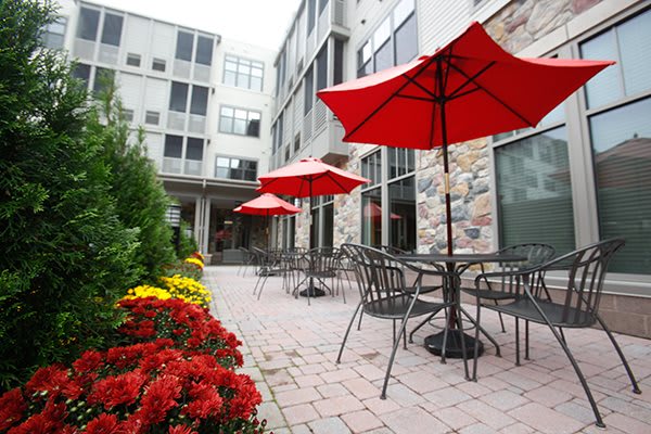 A cozy outdoor patio with red umbrellas, surrounded by flowers and adjacent to a modern building.