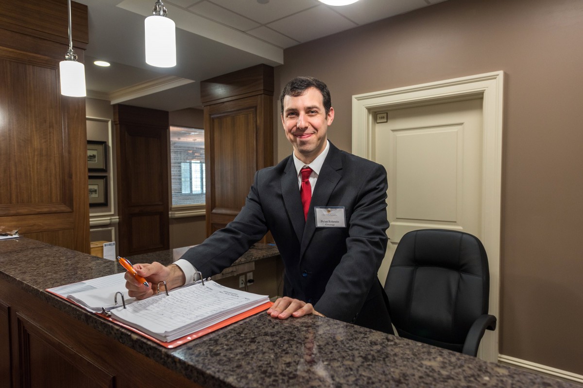 A receptionist standing at a hotel front desk, wearing formal business attire and smiling.