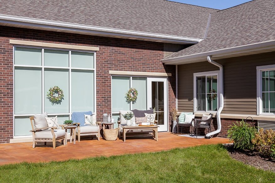 A cozy patio setup with outdoor furniture on a wooden deck attached to a brick house, featuring large windows and decorative wreaths.