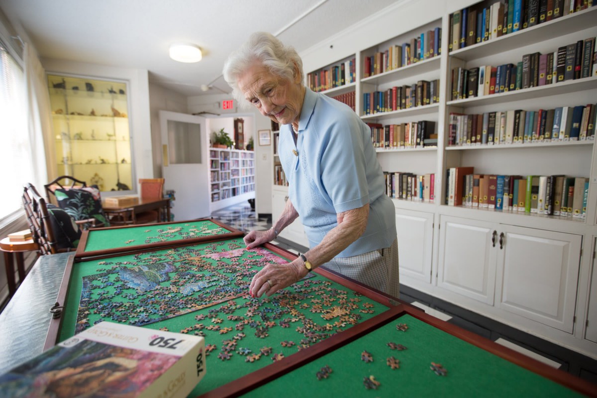 An elderly woman working on a puzzle in a room with bookshelves.