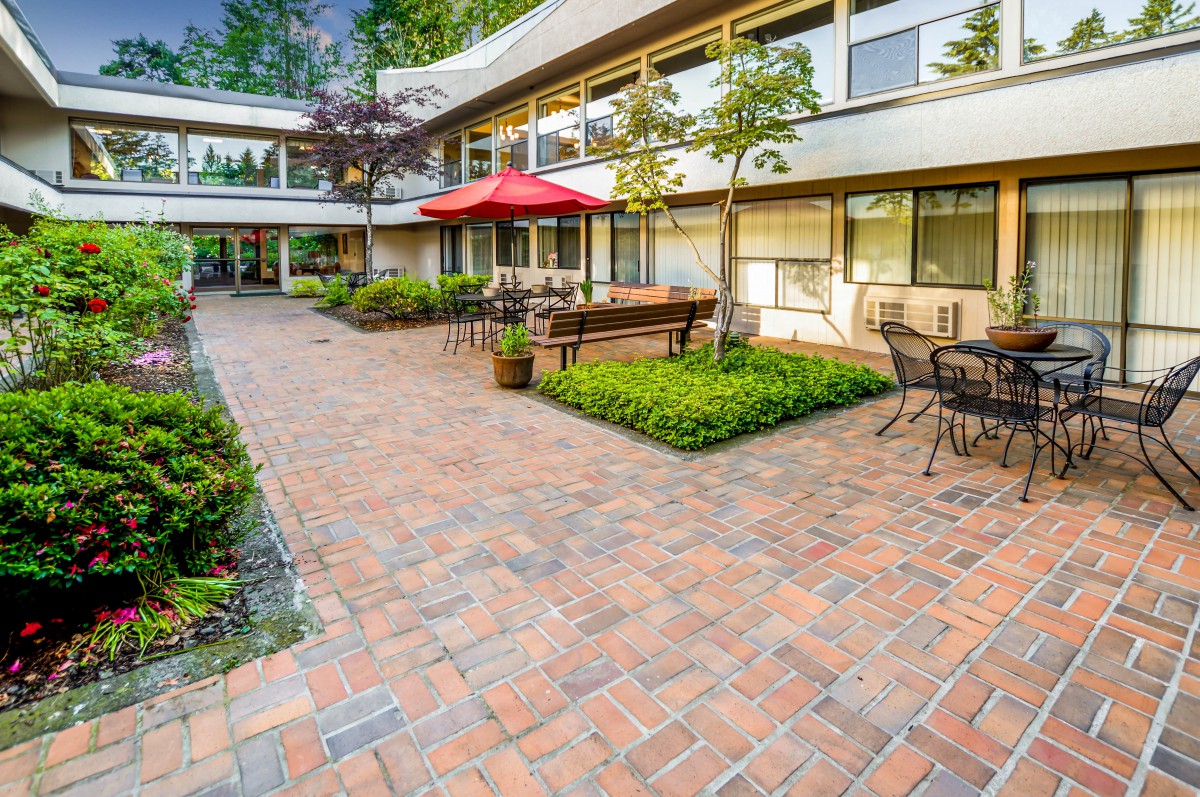 A courtyard with brick paving, outdoor seating, tables, and umbrellas surrounded by greenery.