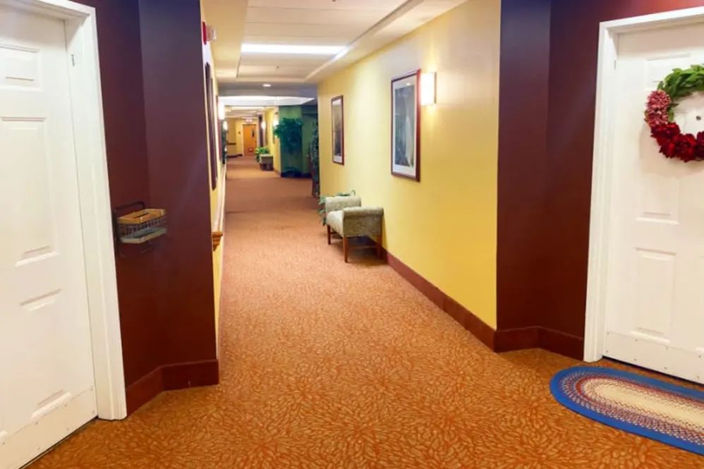 A well-lit hotel hallway with carpeted flooring and a small seating area.