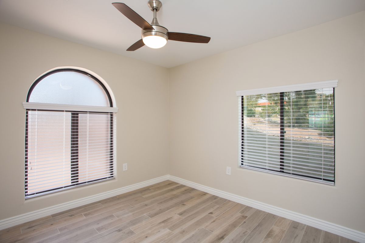 An empty room with a ceiling fan, hardwood flooring, and two windows with blinds.