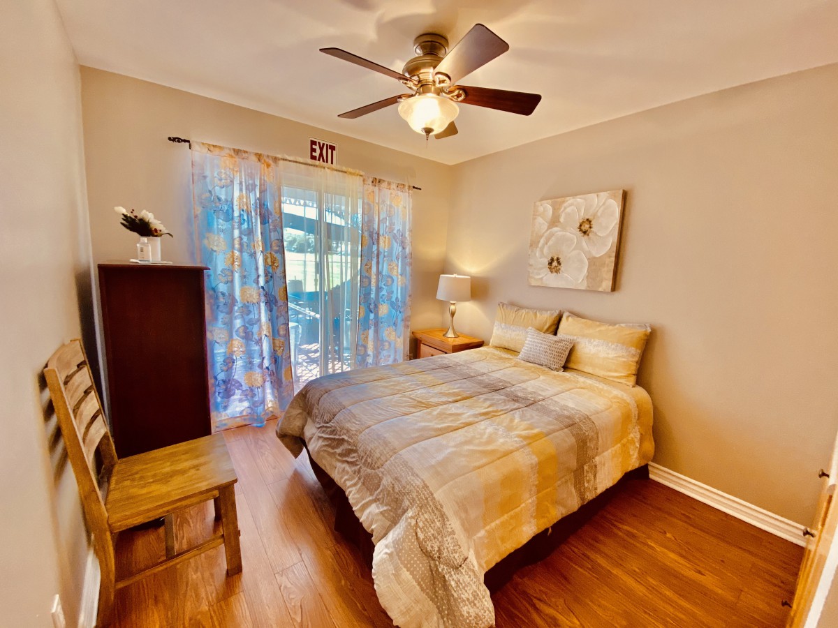 A cozy bedroom with a neatly made bed, ceiling fan, wooden furniture, and soft lighting.