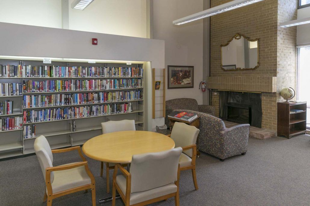 A cozy library reading area with bookshelves, a round table with chairs, and a sofa near a fireplace.