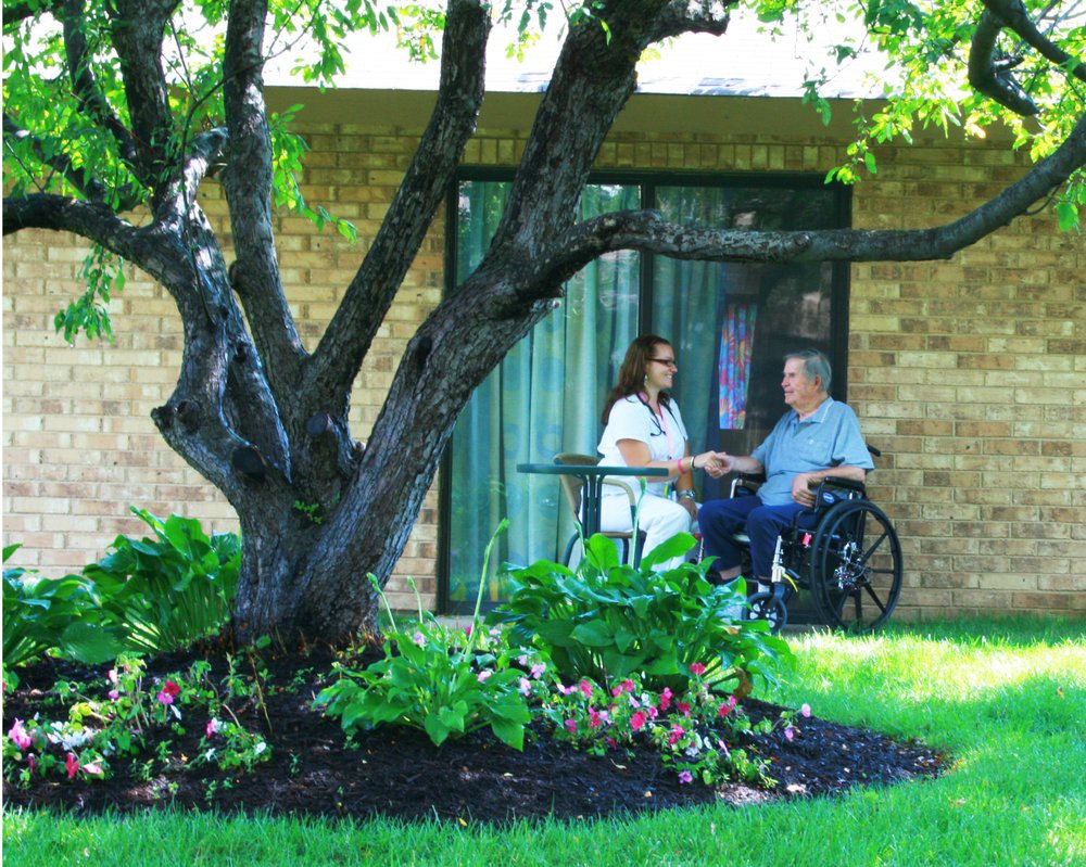 A caregiver assisting an elderly person in a wheelchair outside near a garden.