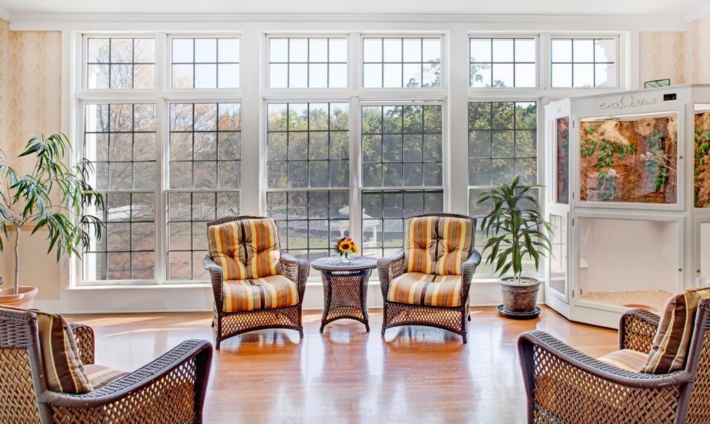 A sunlit room with two cushioned chairs and a small table, surrounded by large windows and a potted plant.