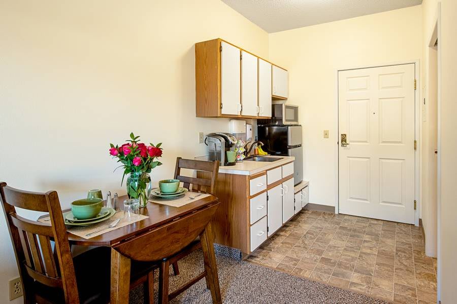 A cozy kitchen with a dining table set for two, featuring green tableware and a bouquet of red flowers.