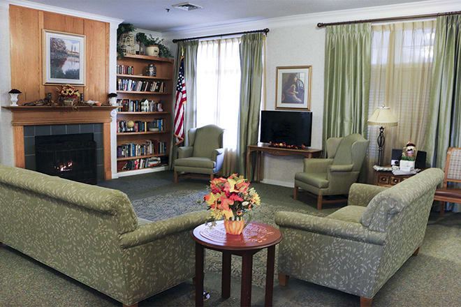 A cozy living room with a seating area, featuring armchairs around a small table with a flower arrangement. The room includes a fireplace, bookshelf, television, and is decorated with curtains.