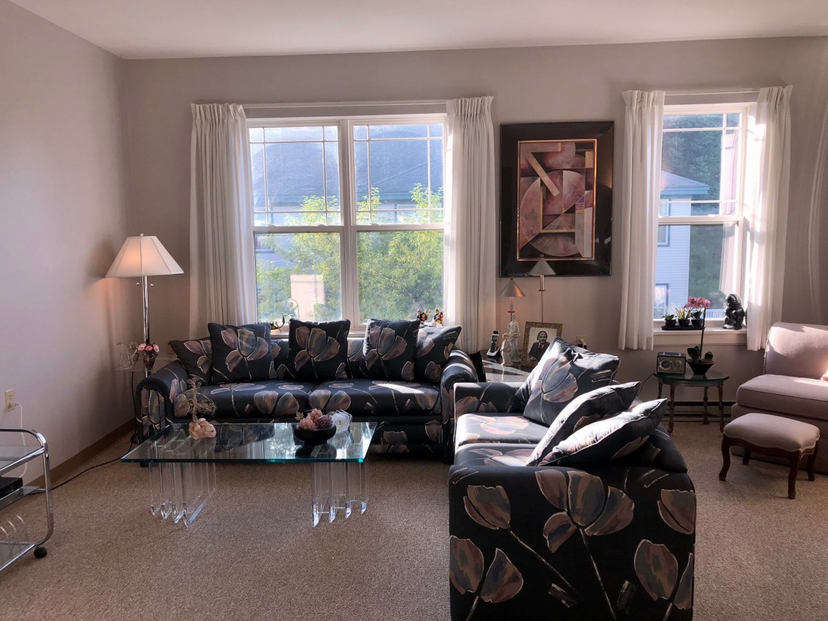 A cozy living room with a patterned sofa and armchair, complemented by a glass coffee table, floor lamp, and large windows with curtains.