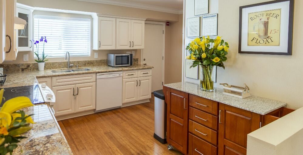 A bright kitchen with white cabinets, hardwood flooring, and a countertop adorned with a vase of yellow flowers.