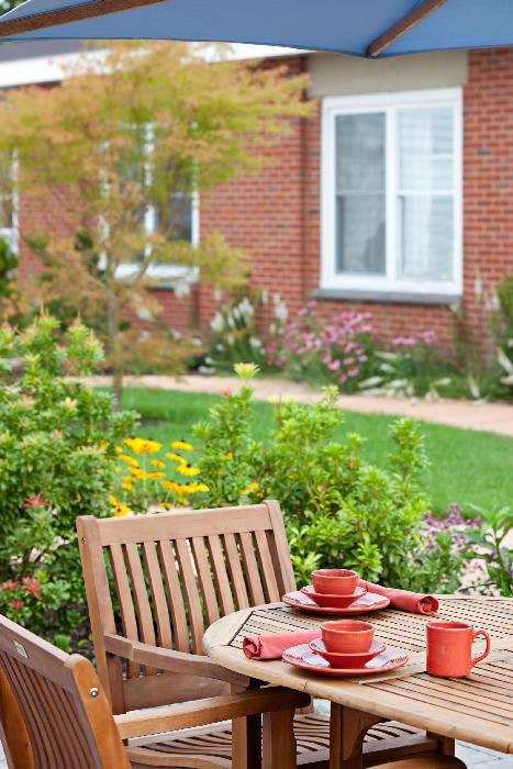 A garden scene with a wooden patio table set for dining, surrounded by vibrant flowers and greenery in front of a brick house.