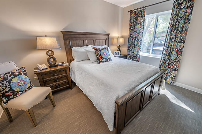 A cozy bedroom with a wooden bed frame, white bedding, and accompanying nightstands with lamps. The room is lit by natural light from a window adorned with floral curtains.