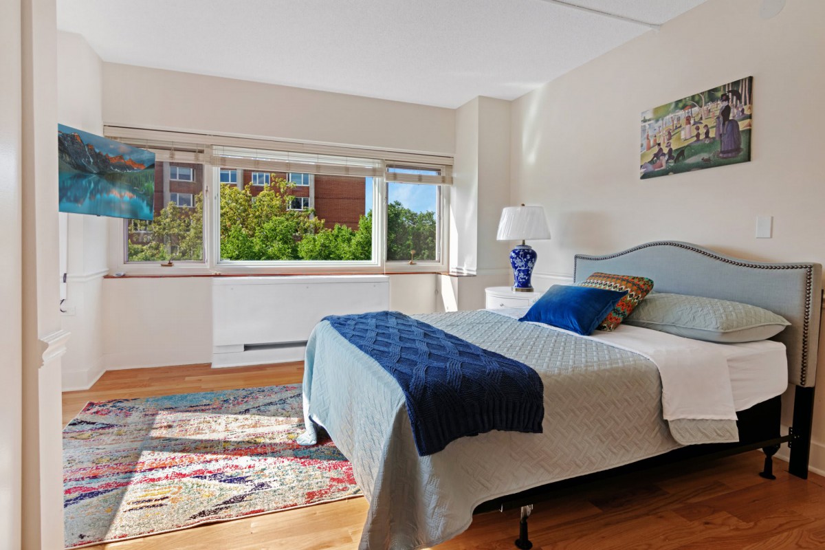 A bright and cozy bedroom with a large window overlooking greenery, featuring a neatly made bed and stylish decor.